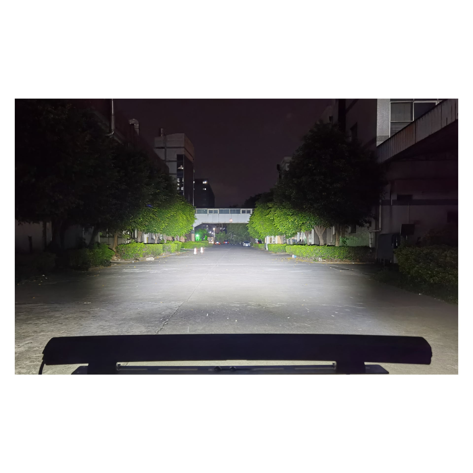 Utsikten från ett fordon på natten visar en starkt upplyst väg framför sig, tack vare Hamron Light bars LED-strålkastare. Träd kantar båda sidor av gatan, och en gångväg förbinder byggnader längre ner på vägen.