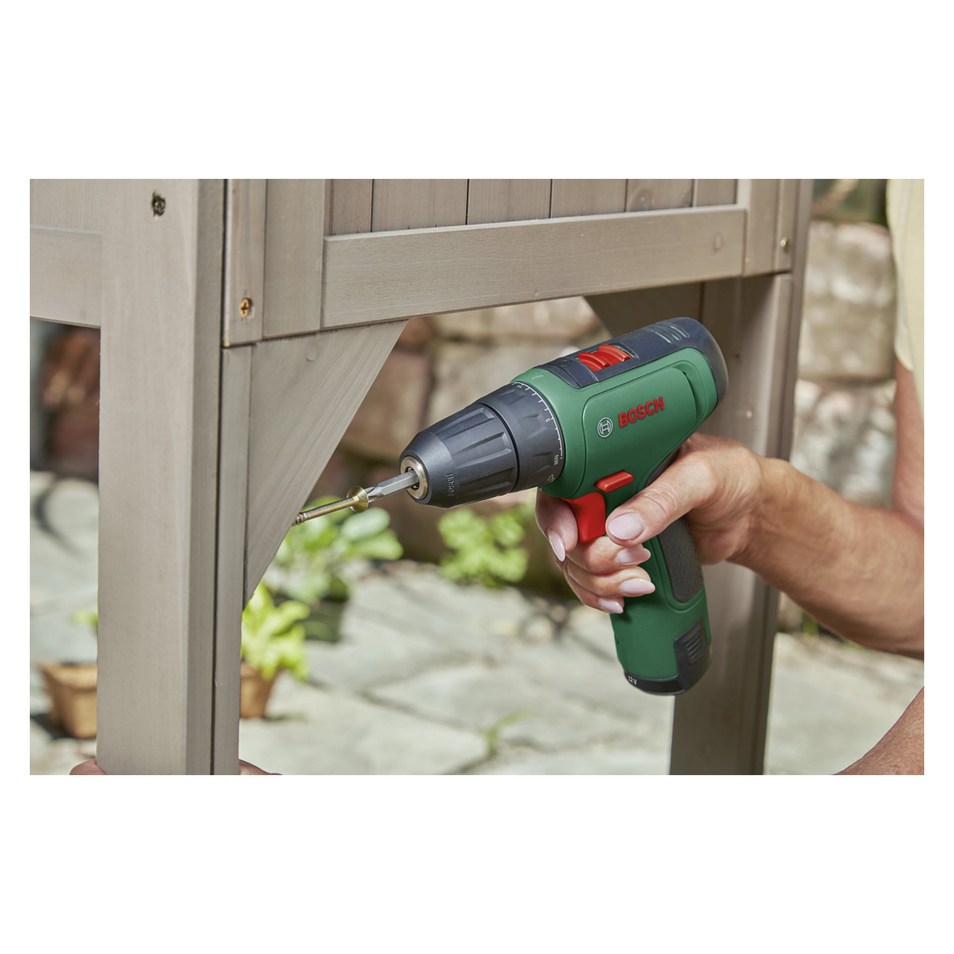 Osoba używa śrubokręta Bosch EasyDrill 1200 Compact z torbą na akcesoria, którą można wkręcić w róg drewnianej konstrukcji na zewnątrz, z ogrodem widocznym w tle.