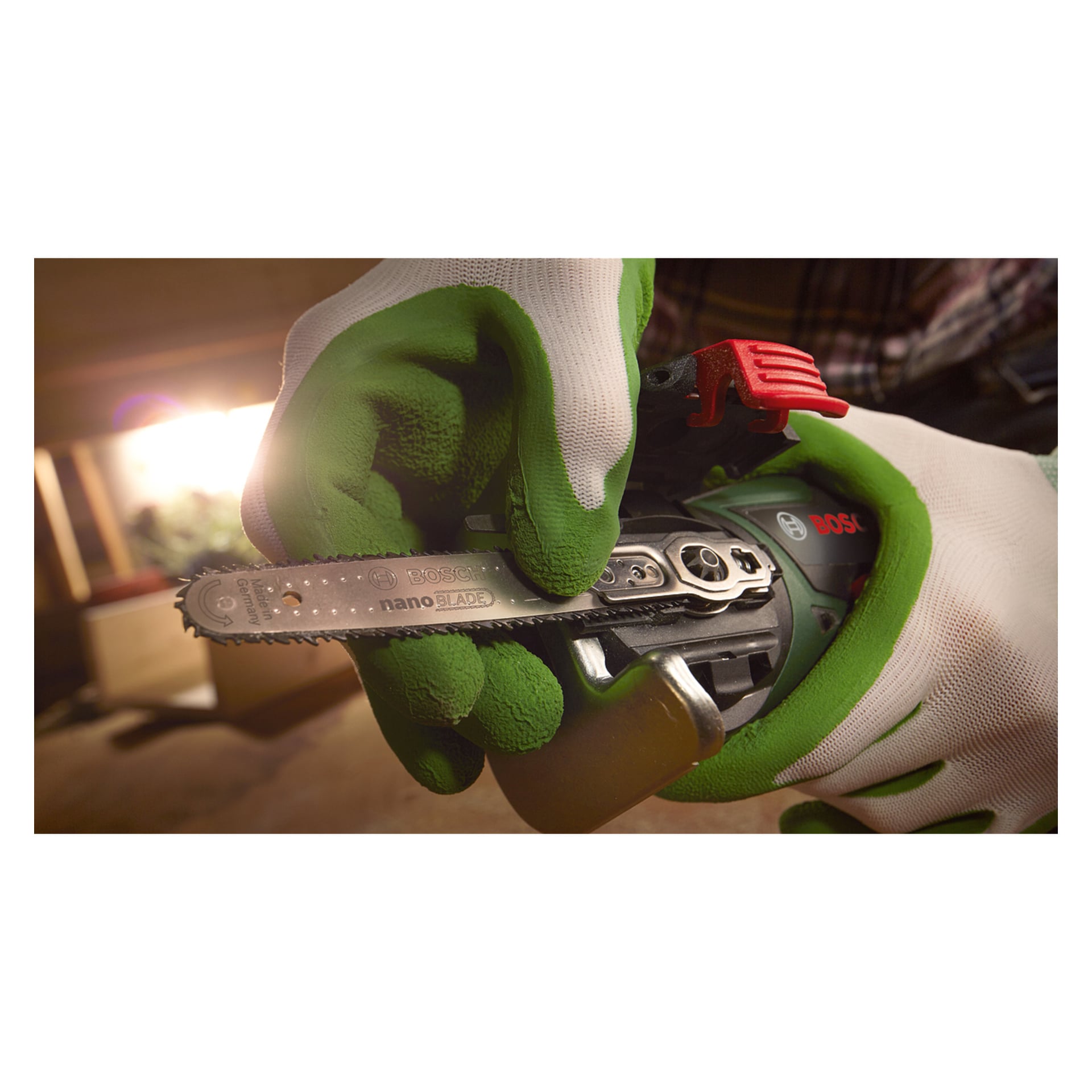 Osoba w zielono-białych rękawiczkach trzyma Bosch Multi-saw EasyCut 12 Solo, z palcem blisko ostrza. Tło jest delikatnie oświetlone i nieostre.