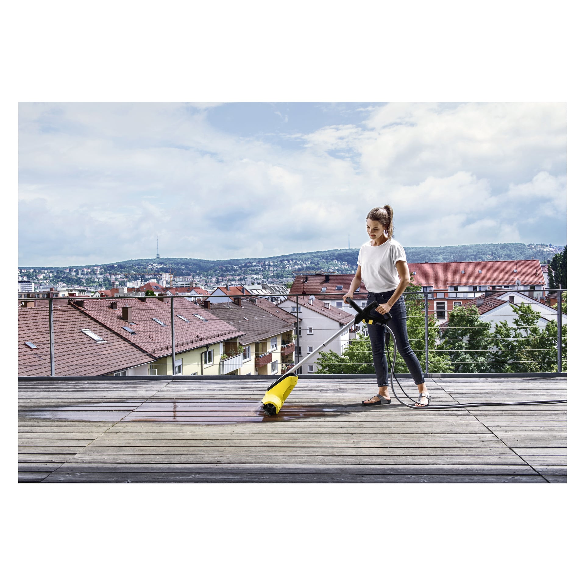 En kvinne iført hvit skjorte rengjør en trebalkongterrasse med den gule Kärcher Terrace wash T-Cleaner PCL4 høytrykksspyleren. I bakgrunnen er det bolighus og åser under en delvis skyet himmel.