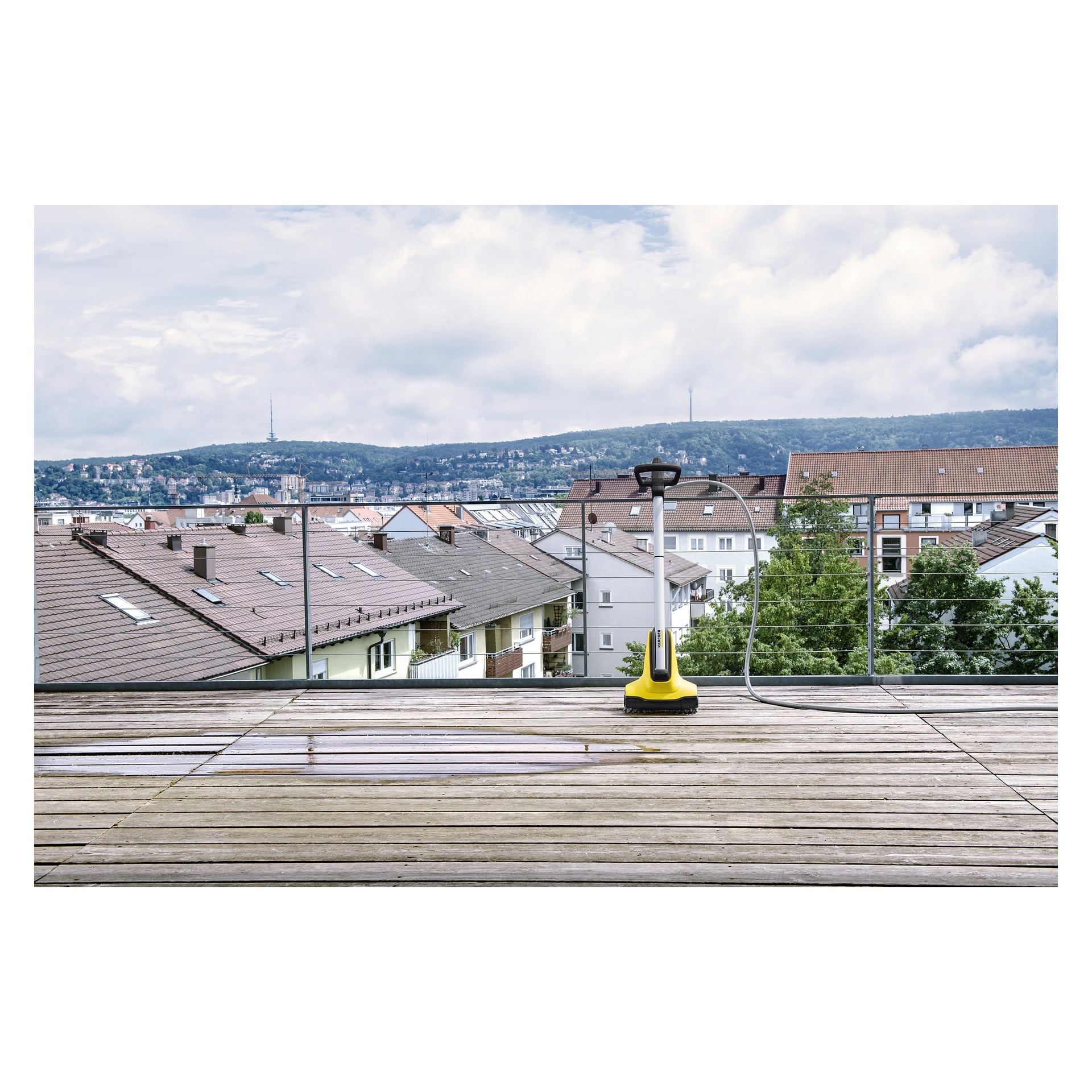 En gul og svart Kärcher Terrace wash T-Cleaner PCL4 hviler på en treterrasse med en merkbar kontrast mellom rengjorte og urensede seksjoner; I bakgrunnen dukker hus og en tredekket ås opp under en skyet himmel.