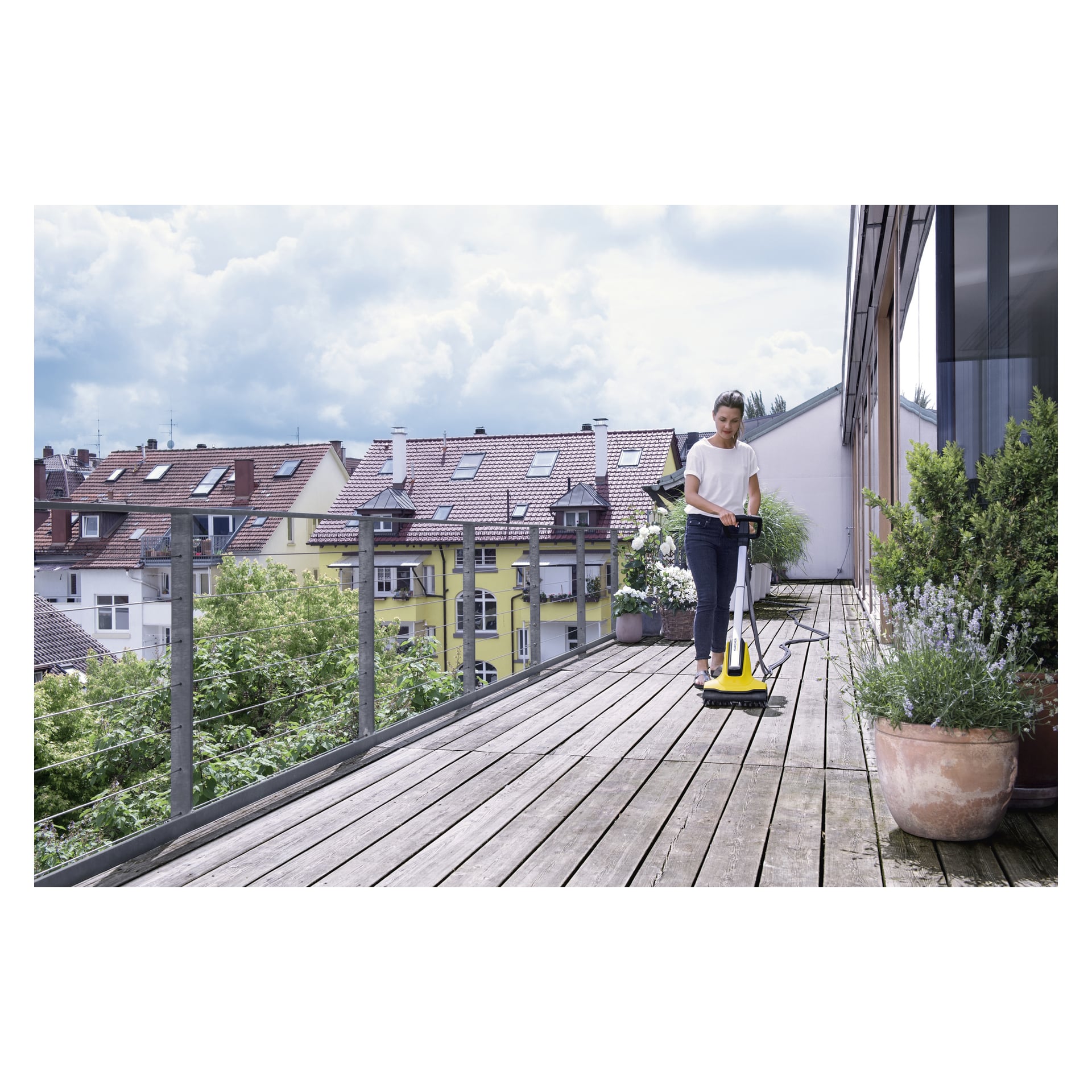 Noen rengjør en treterrasse med den gule Kärcher Terrace-vasken T-Cleaner PCL4 utenfor en moderne bygning, omgitt av potteplanter og med bolighus i det fjerne under en overskyet himmel.