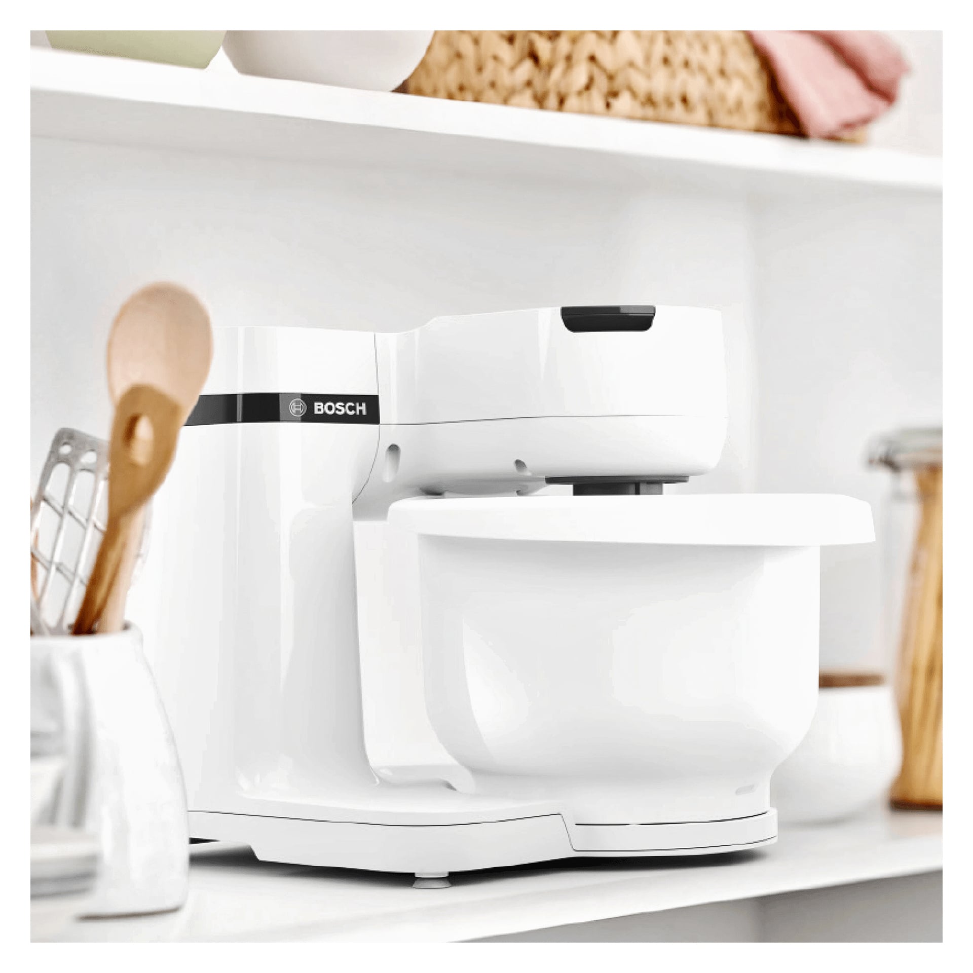 En vit Bosch Home Kitchen MUMS2AW00 apparat står på en bänkskiva, omgiven av trä- och metallredskap, med hyllor och olika köksartiklar i bakgrunden.
