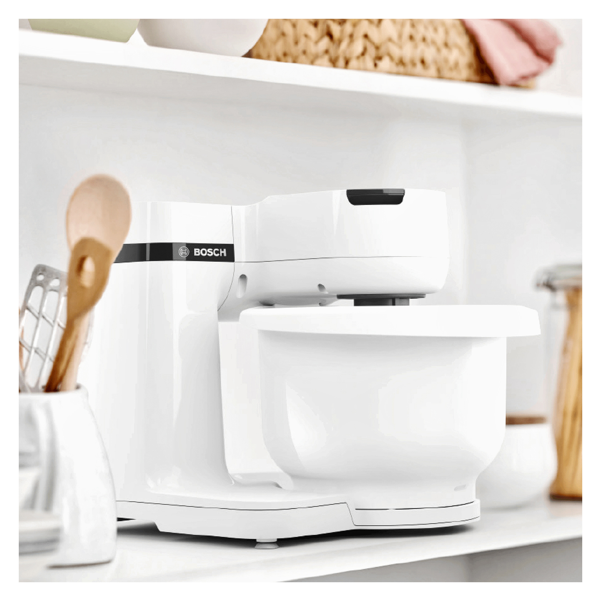En vit Bosch Home Kitchen MUMS2AW00 apparat står på en bänkskiva, omgiven av trä- och metallredskap, med hyllor och olika köksartiklar i bakgrunden.