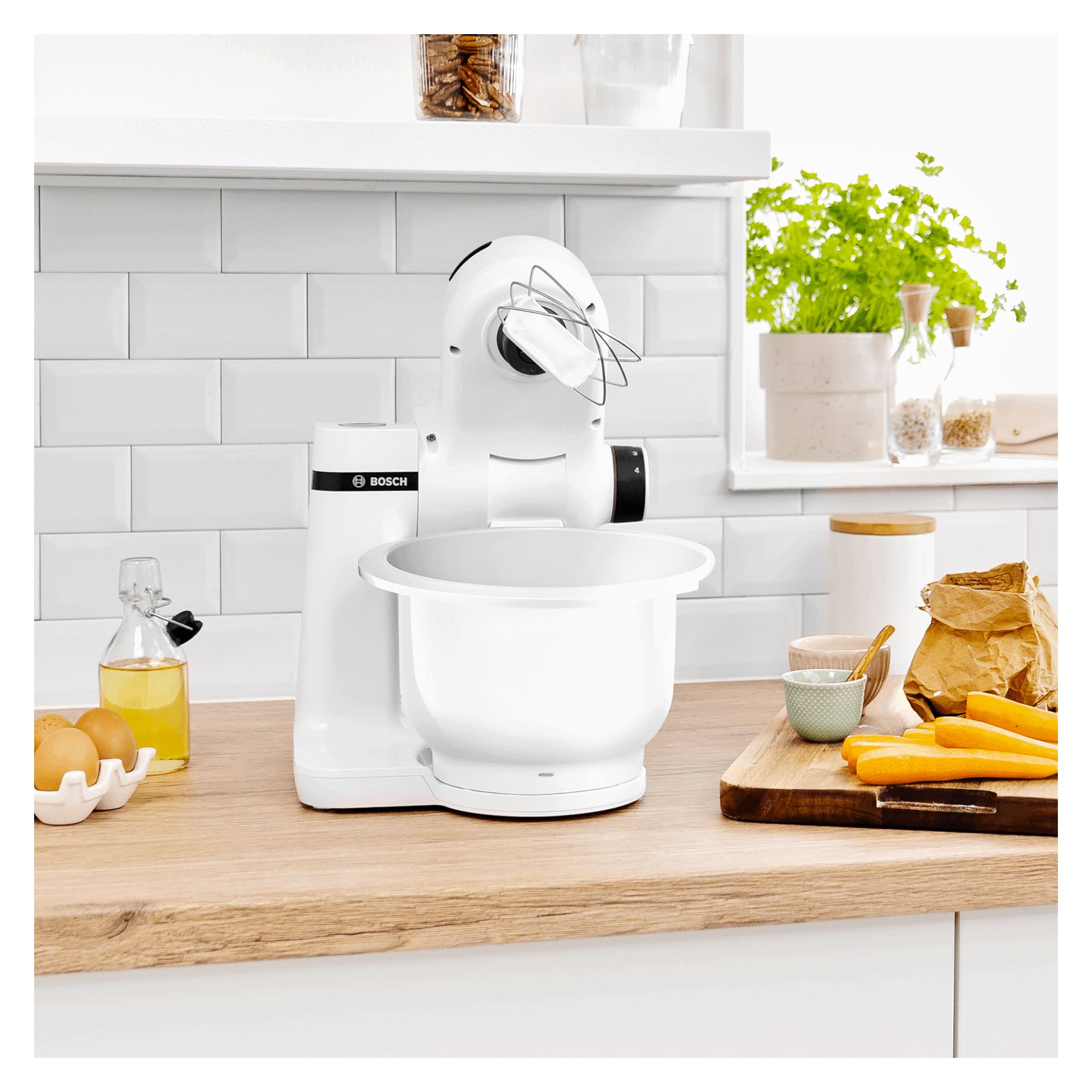 En Bosch köksmaskin modell MUMS2AW00 med vispfäste står på en köksbänk omgiven av ägg, morötter, mjöl och olika andra bakingredienser. Bänken och hyllorna bakom är dekorerade med växter och burkar.