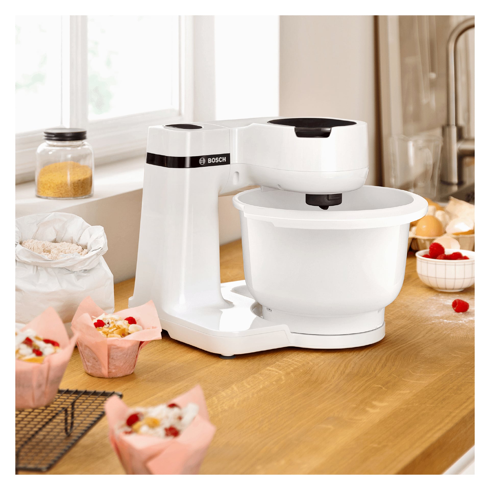 En vit Bosch Home Kitchen-MUMS2AW00 står på en träbänk omgiven av rosa cupcakeförpackningar, en påse mjöl, en sockerburk, ägg och körsbär. Solljuset strömmar in genom fönstret ovanför.