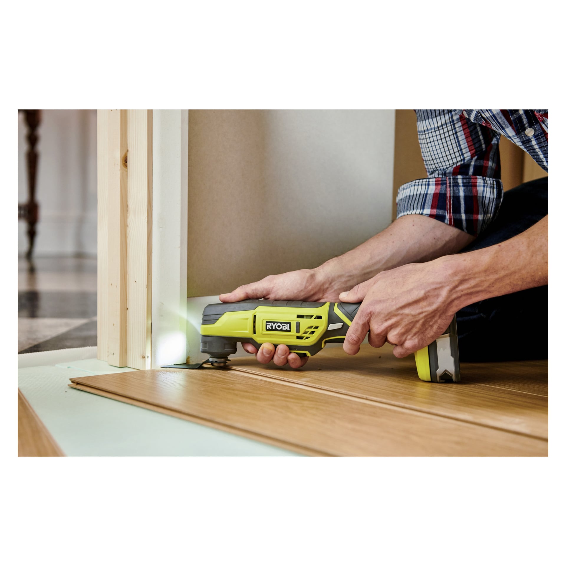 Osoba w koszuli w kratę obsługuje narzędzie Ryobi One+ Multi-tool R18MT3-120GJ, aby przyciąć dolną część framugi drzwi, tworząc miejsce na drewnianą podłogę do przesuwania się pod nią.