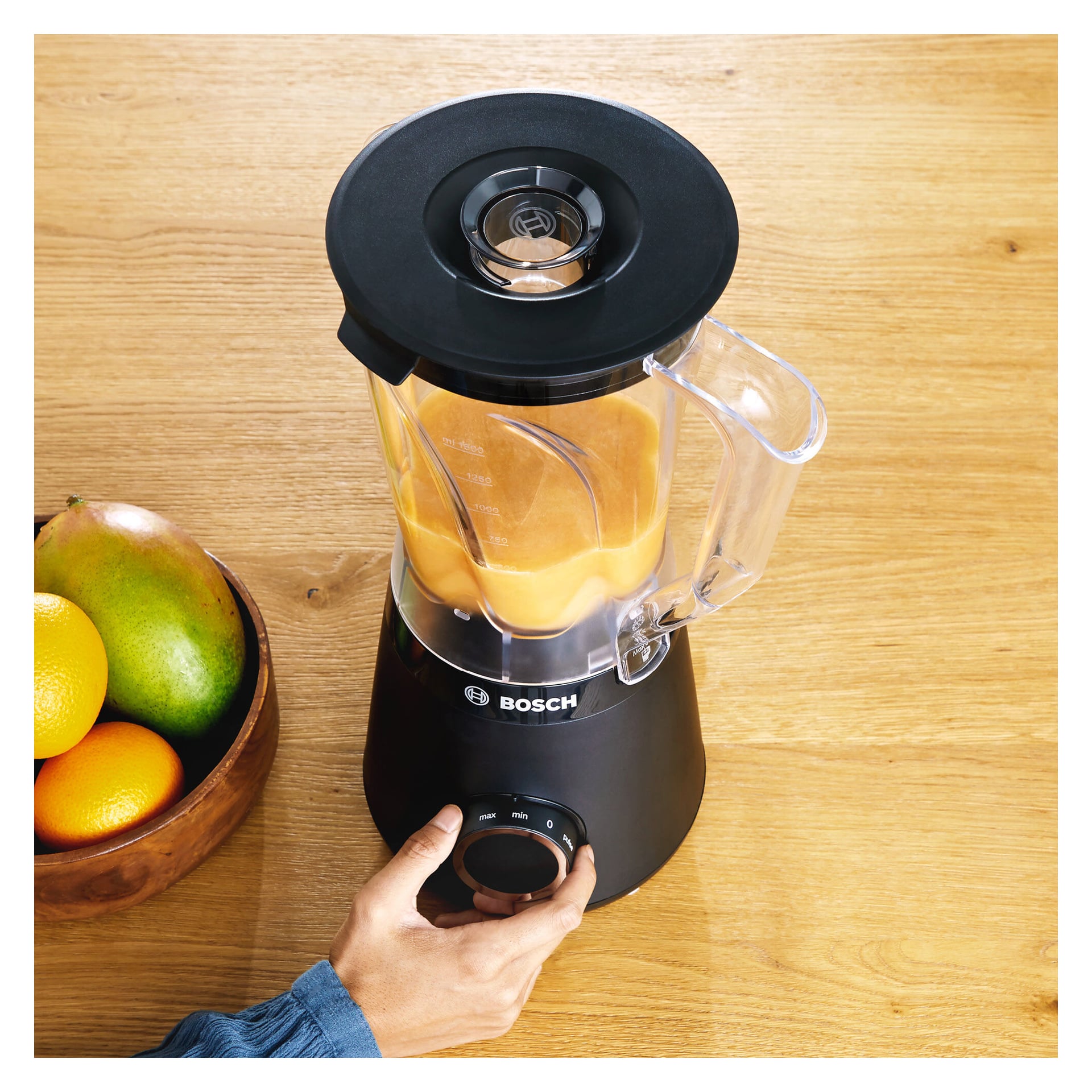 Någon vrider på ratten på en svart Bosch Home Blender MMB6141B innehåller en orange smoothie. Mixern ställs på en träbänk bredvid en skål fylld med mango och apelsiner.