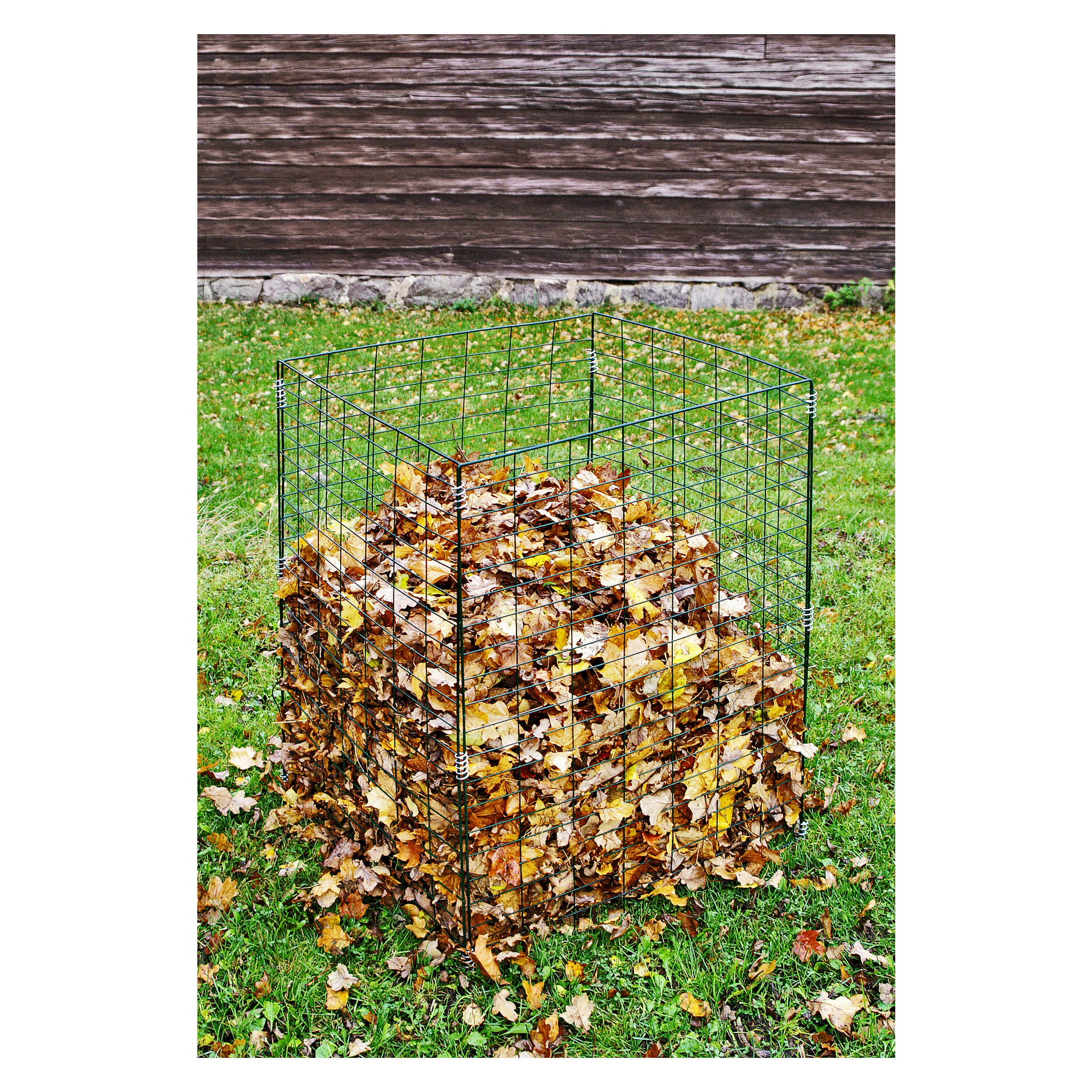 Ett fyrkantigt Hard Head Compost-galler håller en blandning av bruna och gula fallna löv, vilande på en gräsbevuxen gräsmatta. Bakom den står en träbyggnad med horisontell fasad.