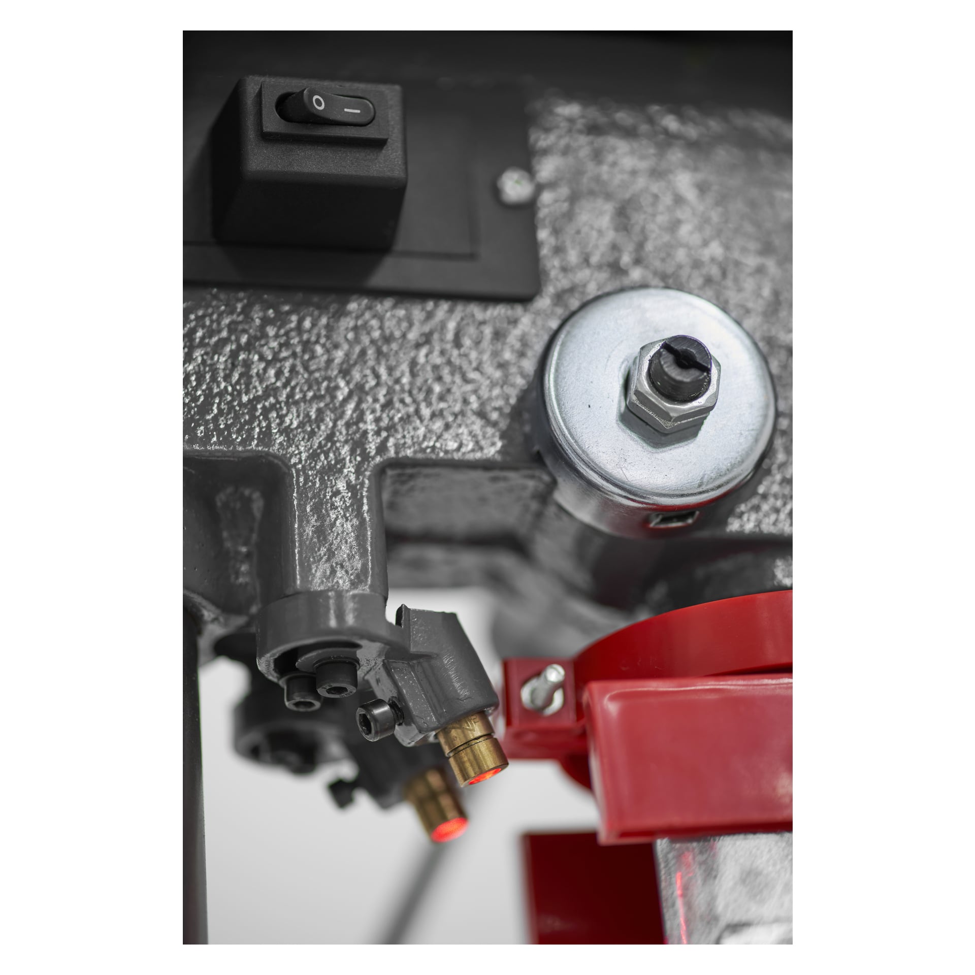 Szczegółowy widok mechanicznych części wiertarki kolumnowej Meec Tools: metalowa obudowa z nakrętką sześciokątną, przełącznik, mosiężne złącza oraz czerwony plastikowy element przymocowany do teksturowanego szarego korpusu.
