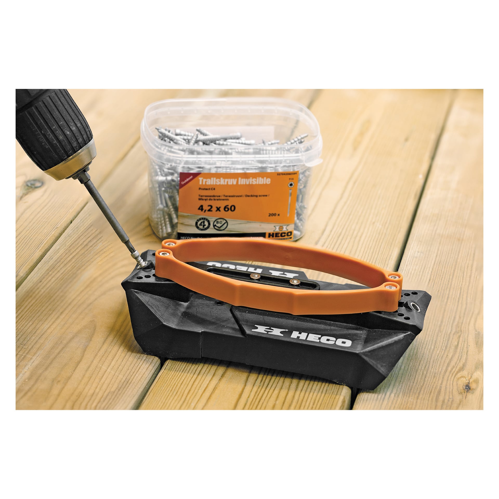 Pokazano bezprzewodową wiertarkę, która wbija śrubę w drewnianą powierzchnię za pomocą czarno-pomarańczowego narzędzia Heco Decking Tool+. Za nim znajduje się plastikowe pudełko wypełnione śrubami HECO Traillskruv Invisible, wszystkie ułożone na drewnie.