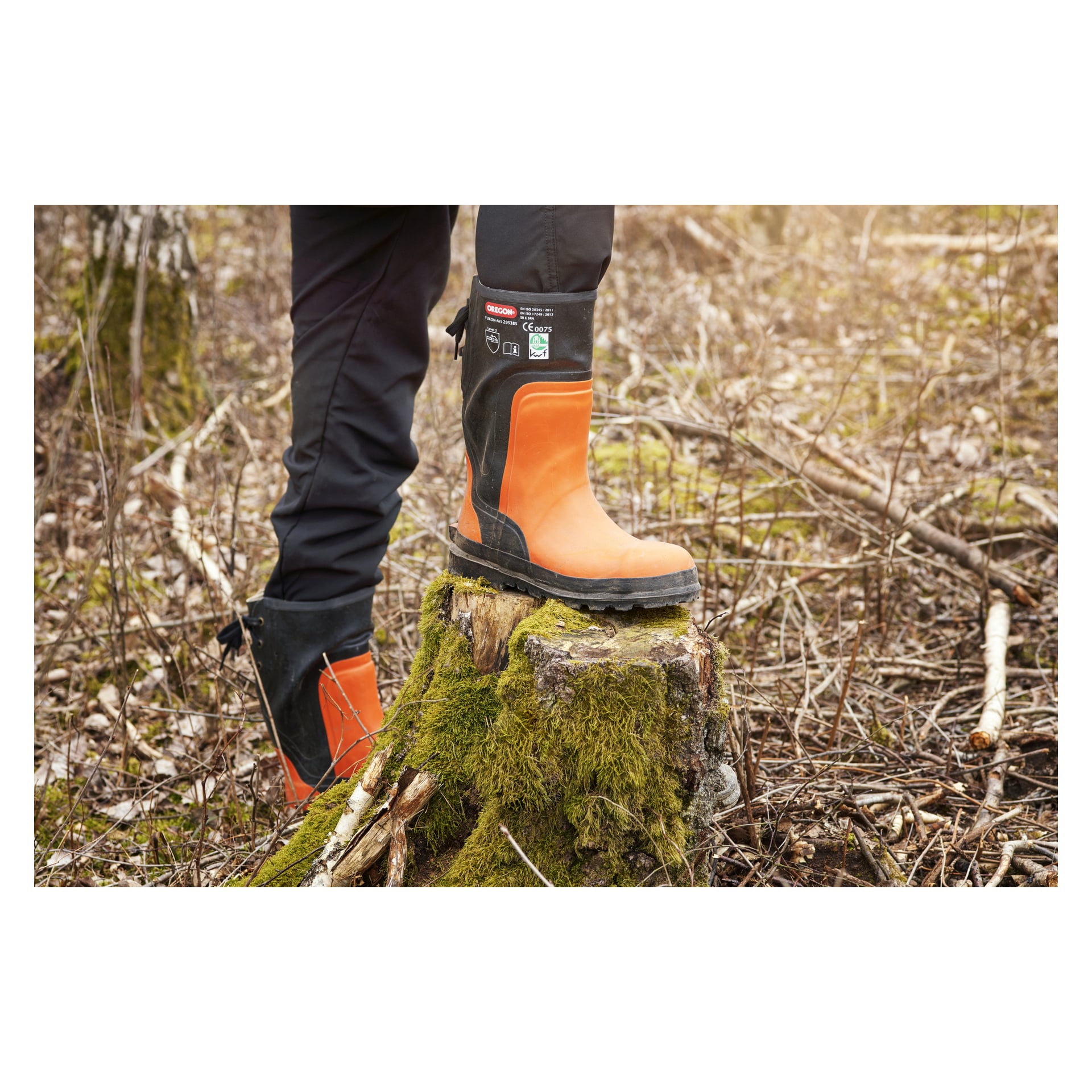 En person i orange och svart Oregon Saw Protection Boots Yukon står i en skogsglänta, vilande en stövel på en mossbeklädd trädstubbe omgiven av torra grenar och löv.