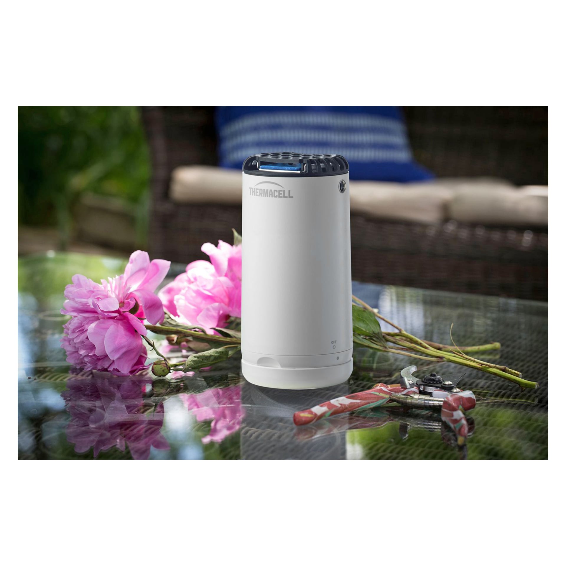En vit myggskyddande Mini Halo från Thermacell står på ett glasbord bredvid rosa blommor och trädgårdssaxar, med en rottingsoffa, en blårandig kudde och grönska i bakgrunden.