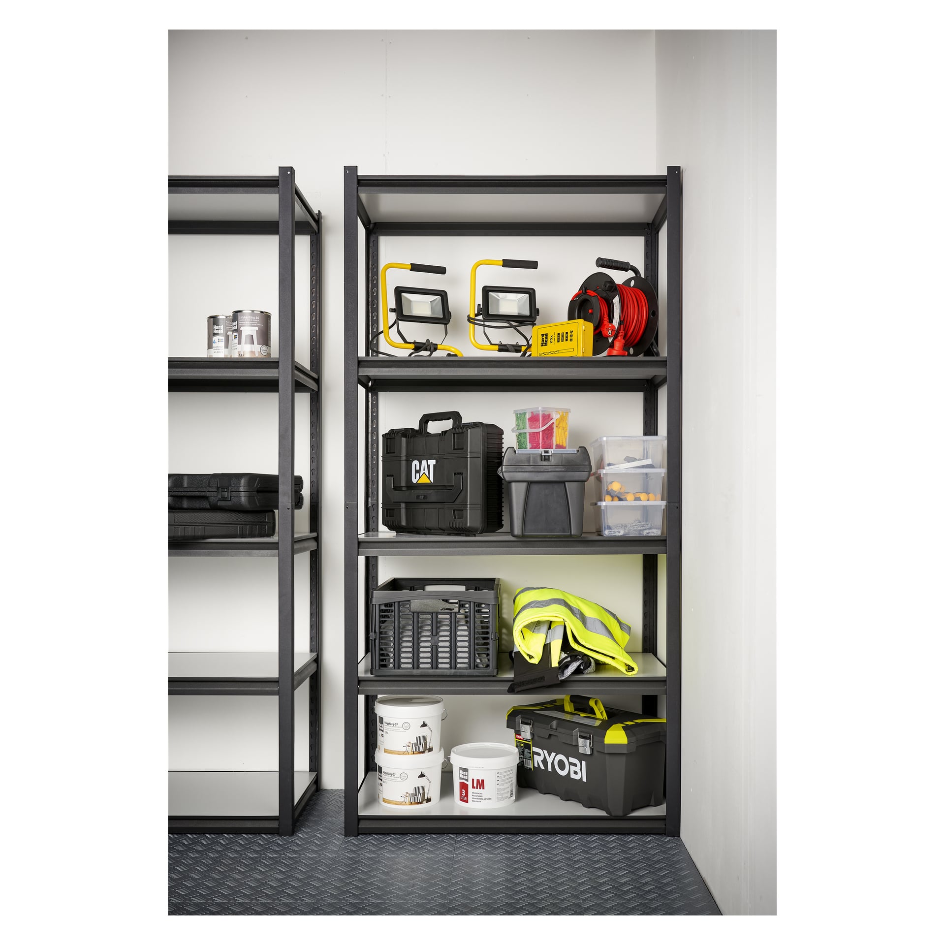 En Hard Head Storage-hylle inneholder arbeidslamper, kabler, en verktøykasse, oppbevaringsbokser, en synlig vest, en Ryobi-koffert, malingsbokser og diverse verktøy og utstyr. Kanten av en annen hylle er delvis synlig på venstre side.