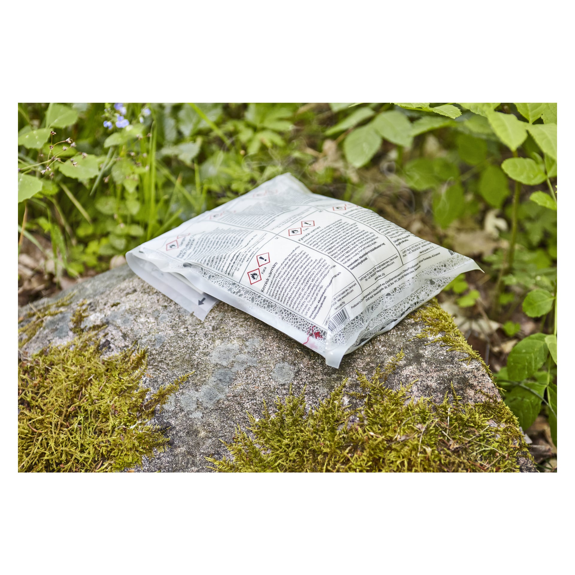 Ett förseglat 24-timmars måltidspaket med värmepaket som visar varningssymboler och instruktioner står ovanpå en mossiga sten utomhus, omgiven av frodiga gröna växter och blad.