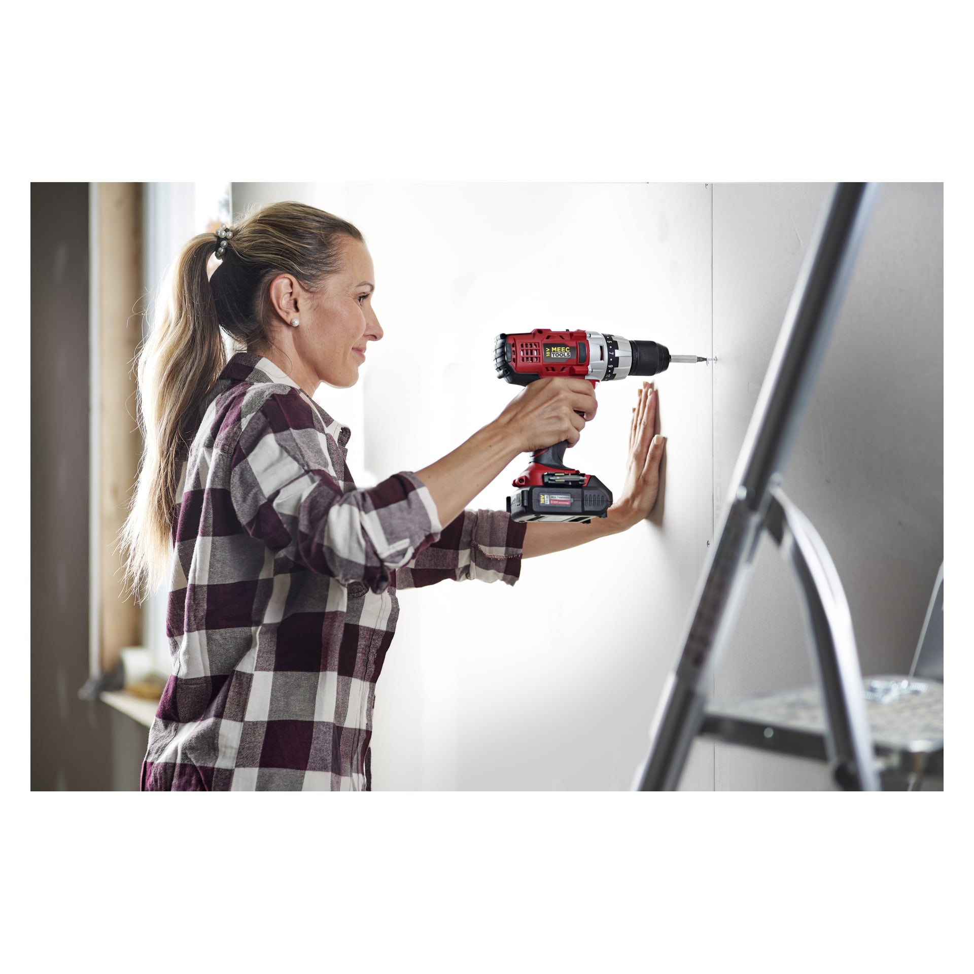 Nainen ruudullisessa paidassa poraa seinään Meec Tools Multiseriesin ruuvimeisselisarjalla DD-18V HOME 500, pitäen poraa toisessa kädessään ja tukemassa seinää toisella; edessä seisoo metallinen tikkaat.