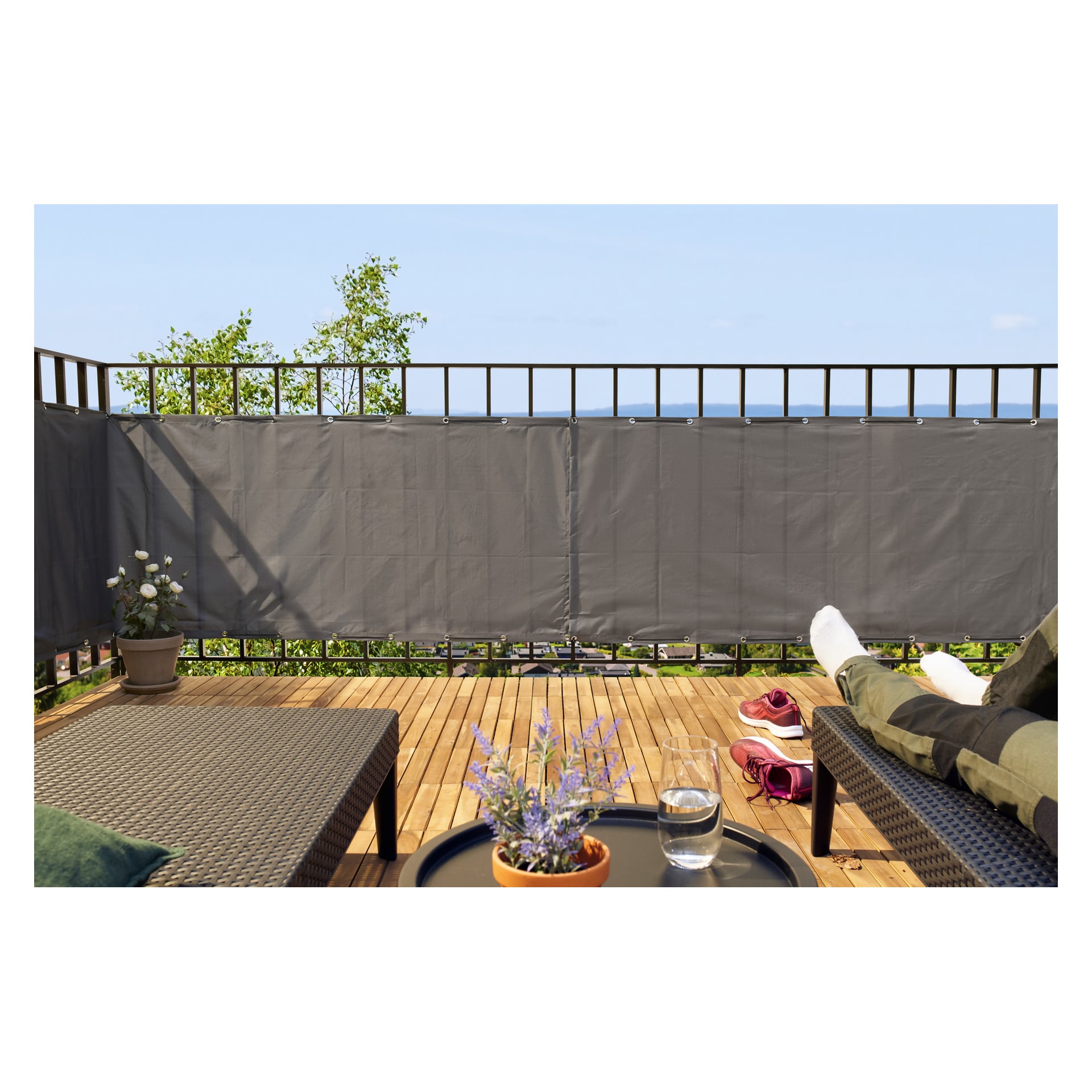 På en träterrass vetter två solstolar mot ett räcke med en Hard Head Balcony insynsskärm. En stol har någons ben utsträckta över sig. Ett litet bord mellan dem har en krukväxt och ett glas vatten, med skor placerade på golvet i närheten.
