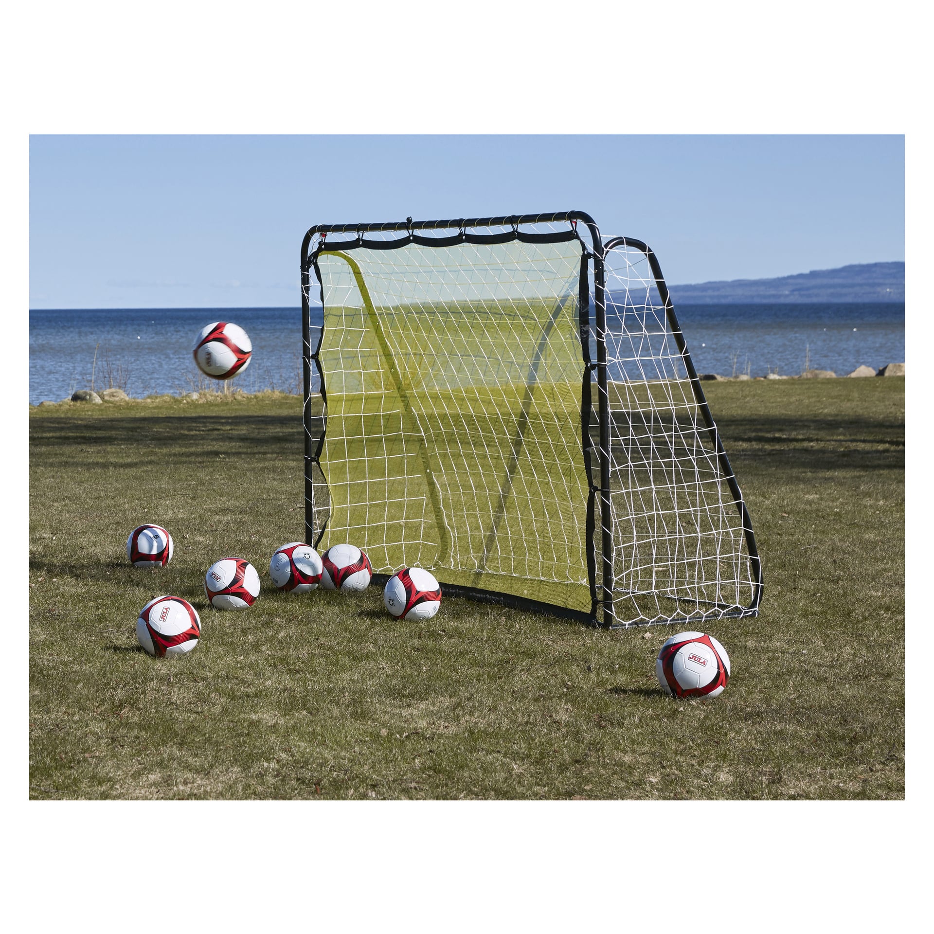 Et Kayoba Football Goal med retur står på gresskledd grunn ved siden av et vann, med flere røde og hvite fotballer spredt rundt og en ball hengende i luften foran målet.