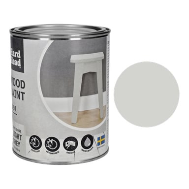 En boks med Hard Head Wood-maling i lys grå med en stor sirkelprøve står ved siden av boksen. Boksen fremhever detaljer og inkluderer et bilde av en krakk malt grå. Teksten på boksen lyder: For innvendig og utvendig treverk, Enkel å påføre, og holdbar finish.