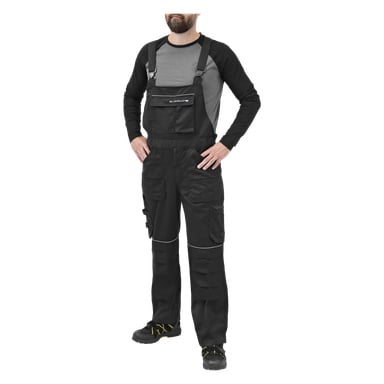 En person står med händerna i sidorna klädd i Bluewear Men's Harry-overalls, som har flera fickor och en framficka med dragkedja. De bär också en grå och svart långärmad skjorta och svarta arbetsskor med gula snören.