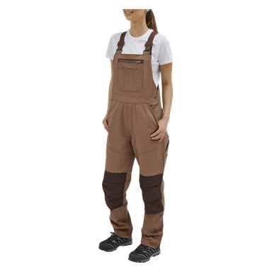 En person kledd i beige Bluewear Women's Overalls Jill med mørkebrune knelapper, en hvit kortermet skjorte og svarte arbeidssko står litt til høyre, hendene hvilende i lommene på kjeledressen.