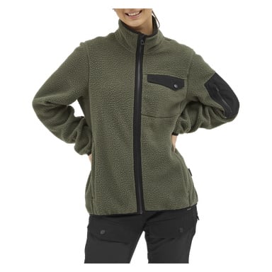 Eine Frau steht mit den Händen in die Hüften gestemmt und trägt eine olivgrüne Kayoba Outdoor Woman's Fleecejacke Ljusdal. Die Jacke verfügt über einen Reißverschluss, eine Brusttasche, die mit einem Druckknopf gesichert ist, und eine schwarze Patch-Tasche am Ärmel. Sie hat ein leichtes Lächeln im Gesicht.