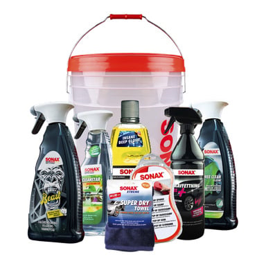 Sonax Car care kit innehåller 8 delar, inklusive rengöringsmedel, sprayer, en handduk och en stor plasthink med rött handtag. Artiklarna är prydligt arrangerade och visar etiketten "Car care kit 8 pieces.".