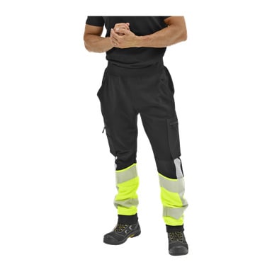 Person som bar svarta Bluewear-byxor med neongula och reflekterande lappar, svart skjorta och stövlar, händerna knäppta; ansiktet syns inte.