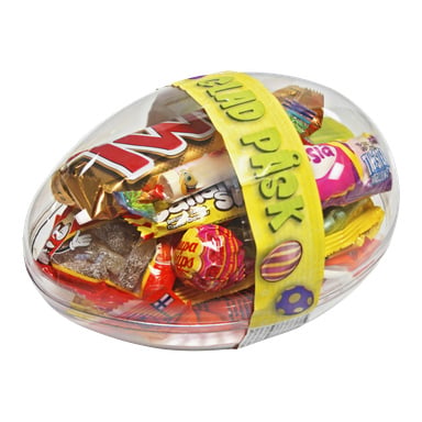 Ett genomskinligt plastägg innehåller diverse godis och choklad, inslaget med ett gult band som det står GLAD PÅSK på och har påskäggsillustrationer.