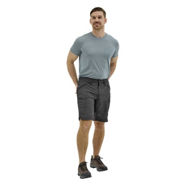 En man står framåt i en ljusblå T-shirt, mörkgrå cargoshorts och bruna vandringsskor, ena handen bakom ryggen, uttrycket neutralt.