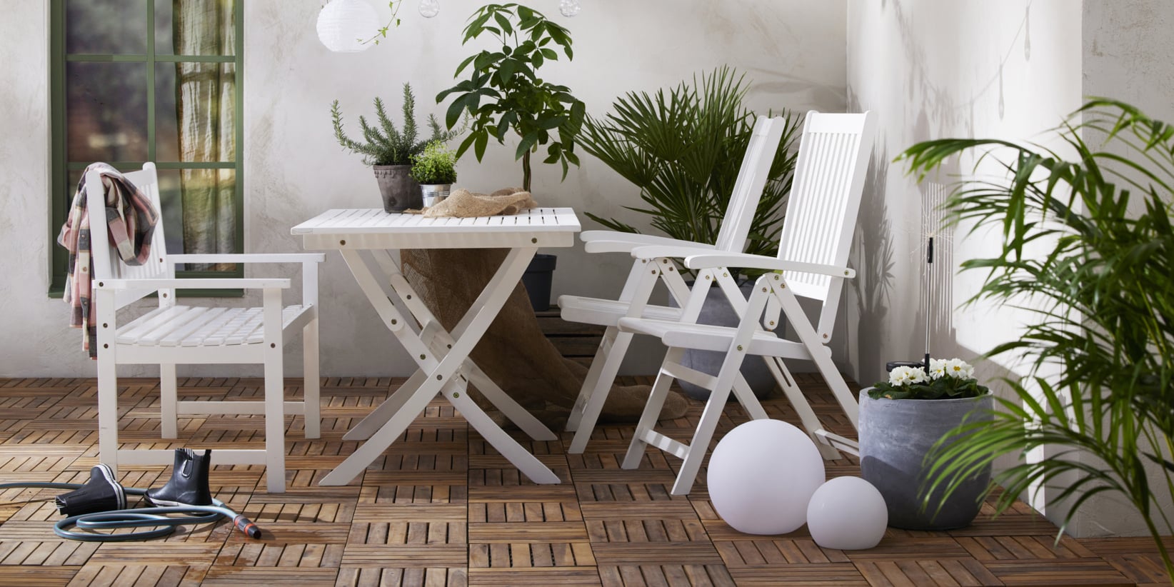 Eine gemütliche Terrasse mit einem weißen Tisch und zwei Stühlen auf gemustertem Holz, umgeben von Topfpflanzen, Schuhen, einem Schlauch und zwei weißen Kugeln.