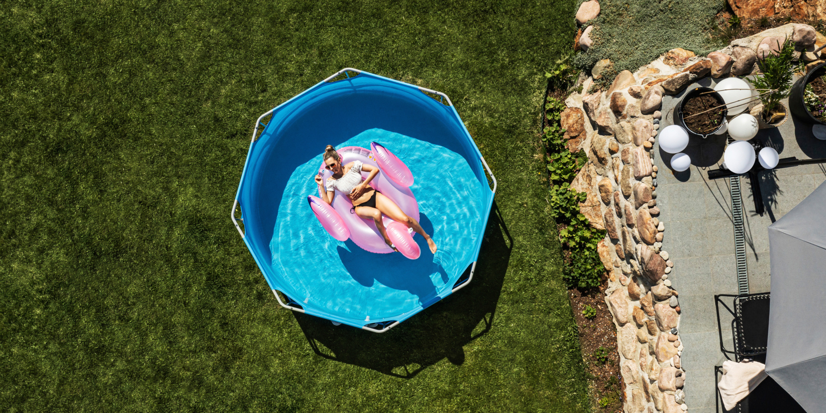 Eine Person liegt auf einem rosa Poolschwimmer in einem kleinen blauen Pool auf grünem Gras, neben dem von oben gesehen Terrassensteine und Topfpflanzen stehen.