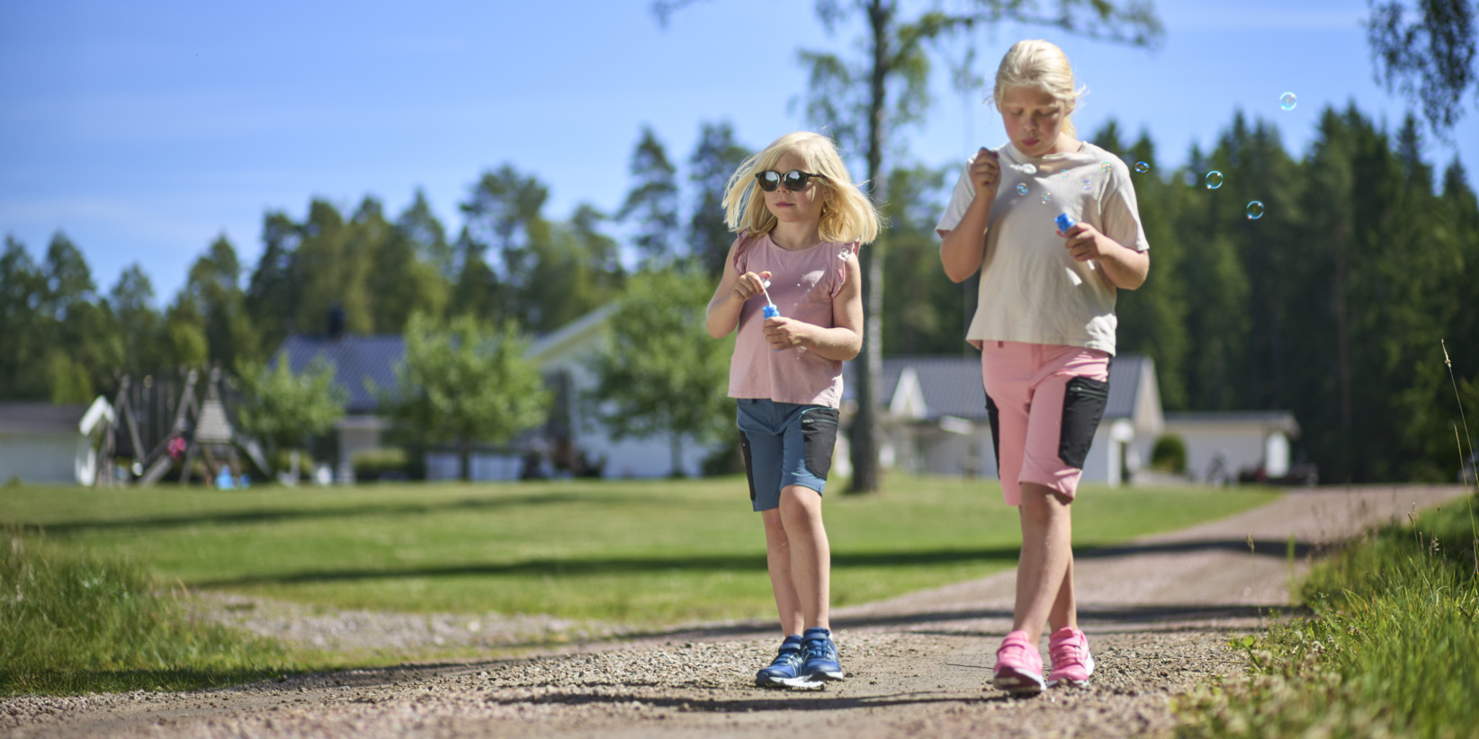 En solig dag promenerar två barn längs en grusväg och blåser såpbubblor. Träd, gräs, hus bakom; Man bär solglasögon.