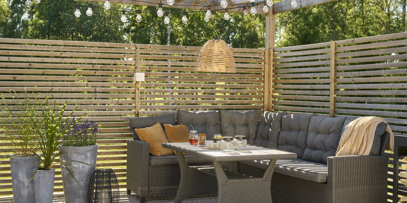 Moderne terrasse med et grått rattansofasett, bord, puter, pledd, potteplanter, kurvlampe, lyslenker og inngjerdet privatliv.