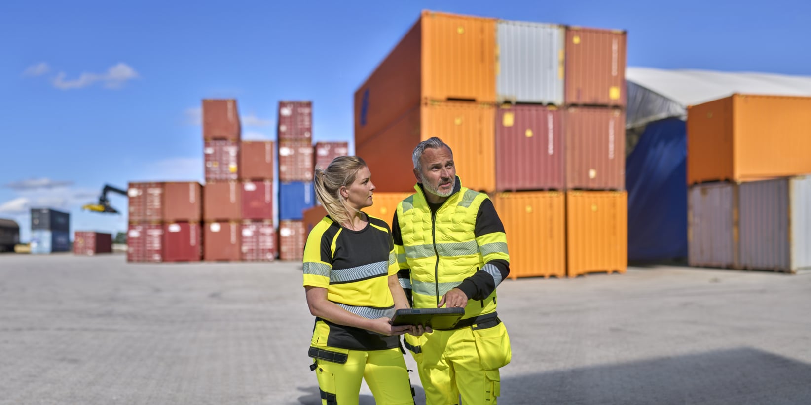 To arbeidere i advarselsklær står på et skipsverft og holder et brett i hendene. I bakgrunnen ser vi stablede containere og en kran, og himmelen er blå.