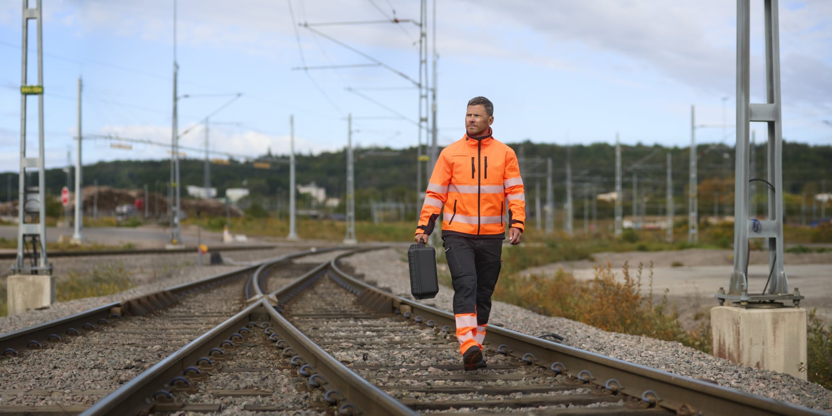 Osoba ubrana w pomarańczową kamizelkę odblaskową i spodnie idzie wzdłuż torów kolejowych, niosąc czarną walizkę. Linie energetyczne i metalowe konstrukcje otaczają tory, a tłem są drzewa i niebo.