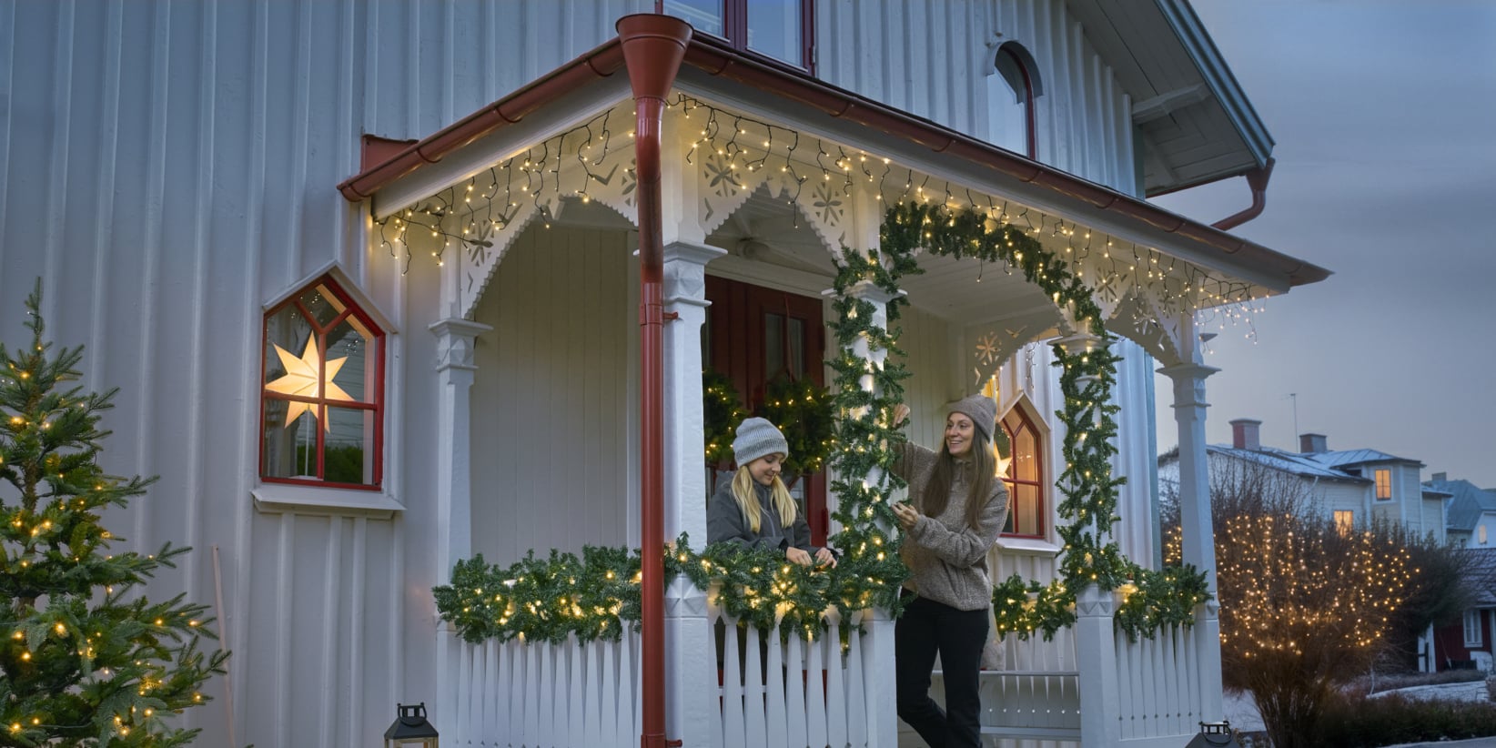 To personer i vinterfrakker prater på en veranda til et hvitt hus utsmykket med lyslenker, kranser og glødende juletrær i skumringen.