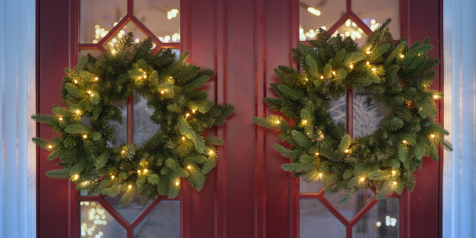 Zwei grüne Kränze mit warmweißen Lichtern schmücken rote Flügeltüren, während im Hintergrund sanfte, verschwommene Weihnachtslichter leuchten.