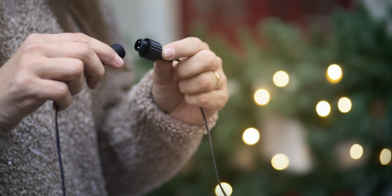 En persons händer förenar två svarta utomhusljusslingor, med festliga suddiga lampor och grönt lövverk mjukt i bakgrunden.
