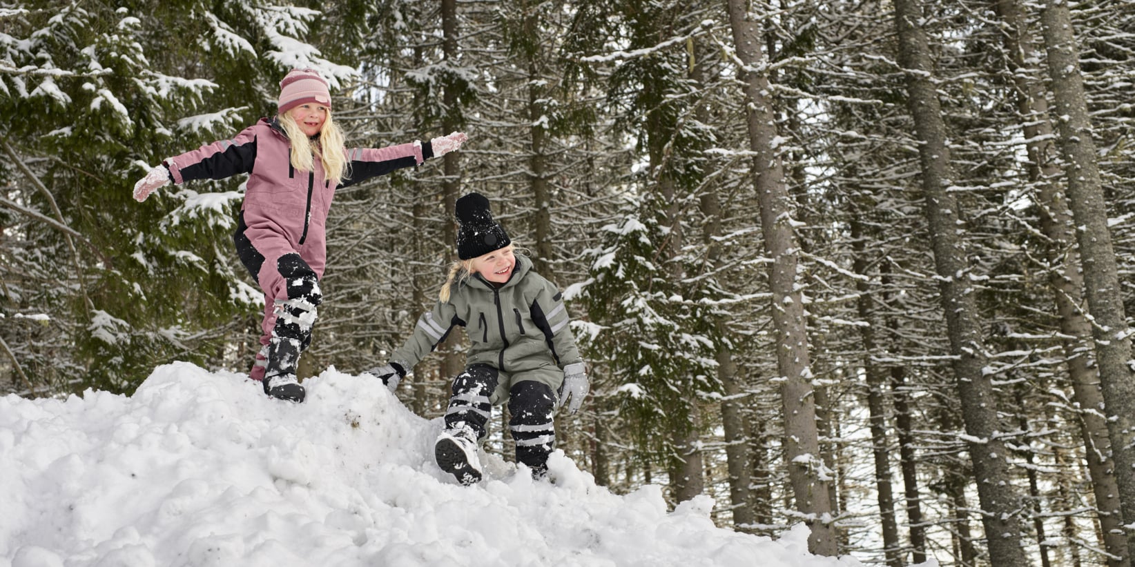 Två barn i vinterkläder leker på en snöig kulle i en skog, omgiven av snöpudrade träd. Den ene står medan den andre hukar sig, båda ler.