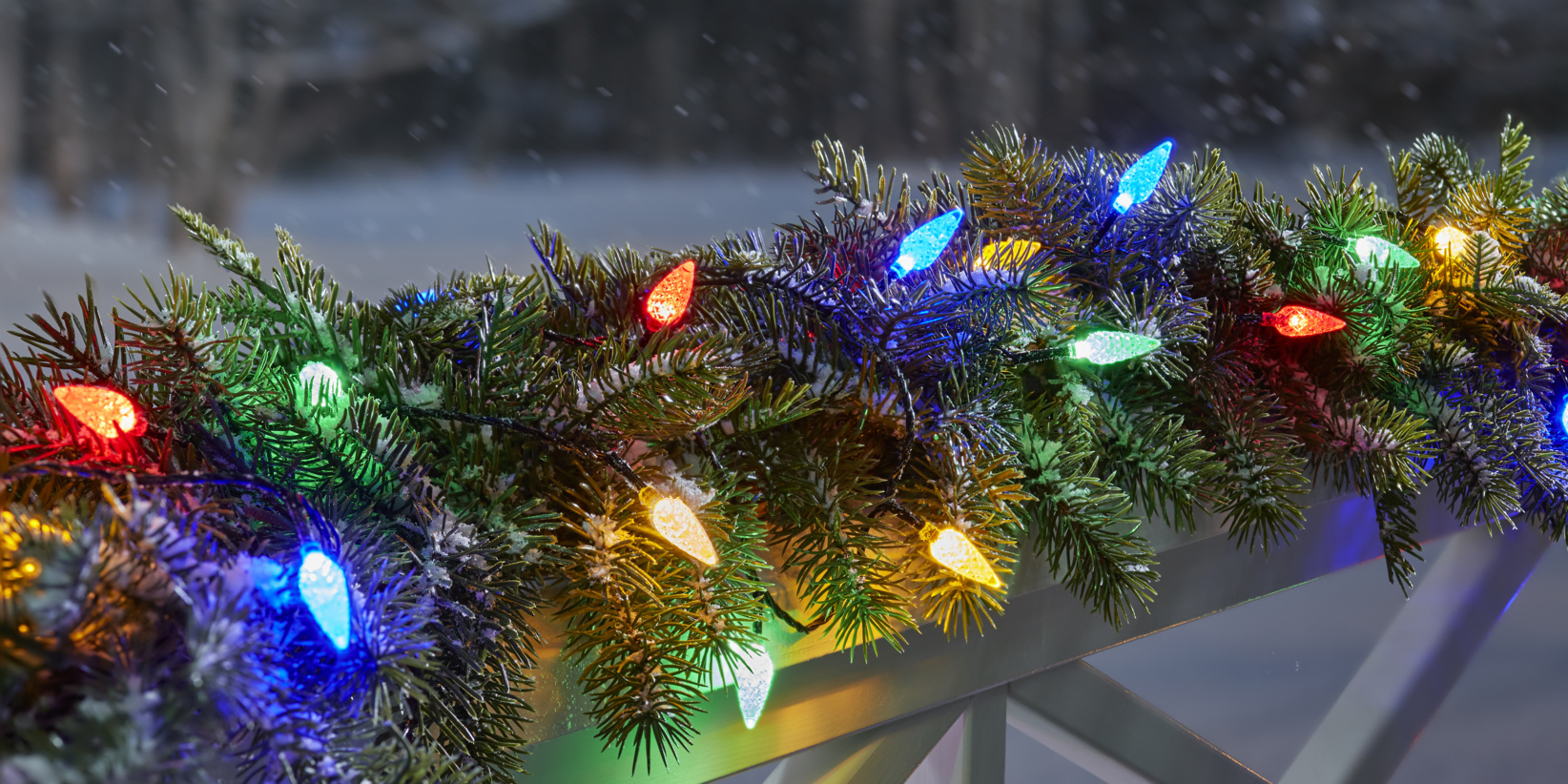 Kieferngirlande mit großen, bunten roten, blauen, gelben, grünen und weißen Weihnachtslichtern, die über einem Geländer mit Schnee dahinter drapiert sind.