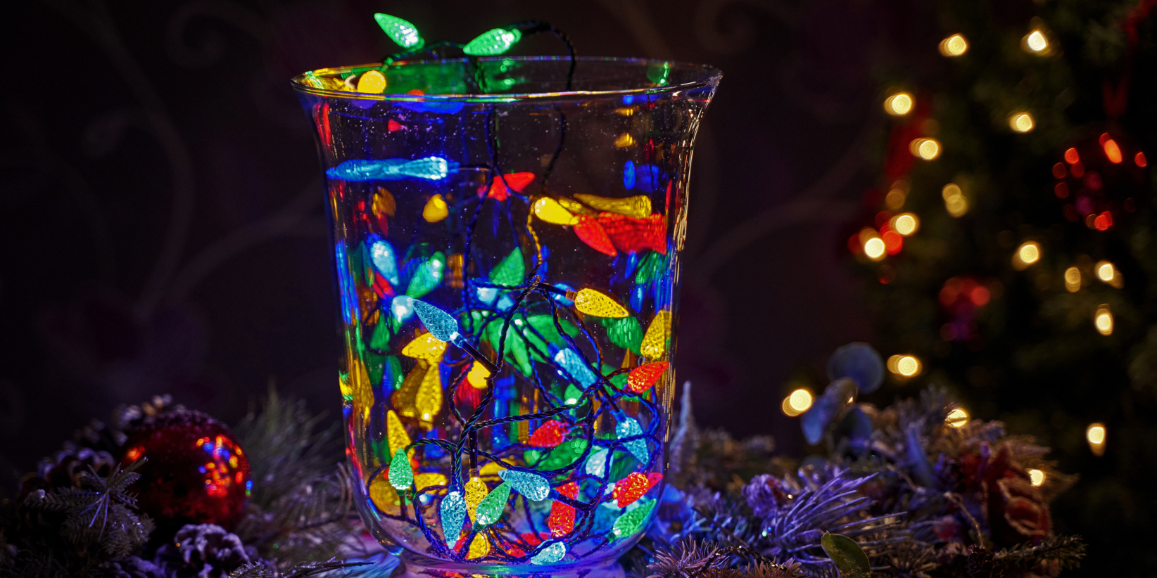 In einer Glasvase stehen bunte LED-Lichter in Blau, Grün, Gelb und Rot, umgeben von Tannenzweigen und verschwommener Weihnachtsbeleuchtung.