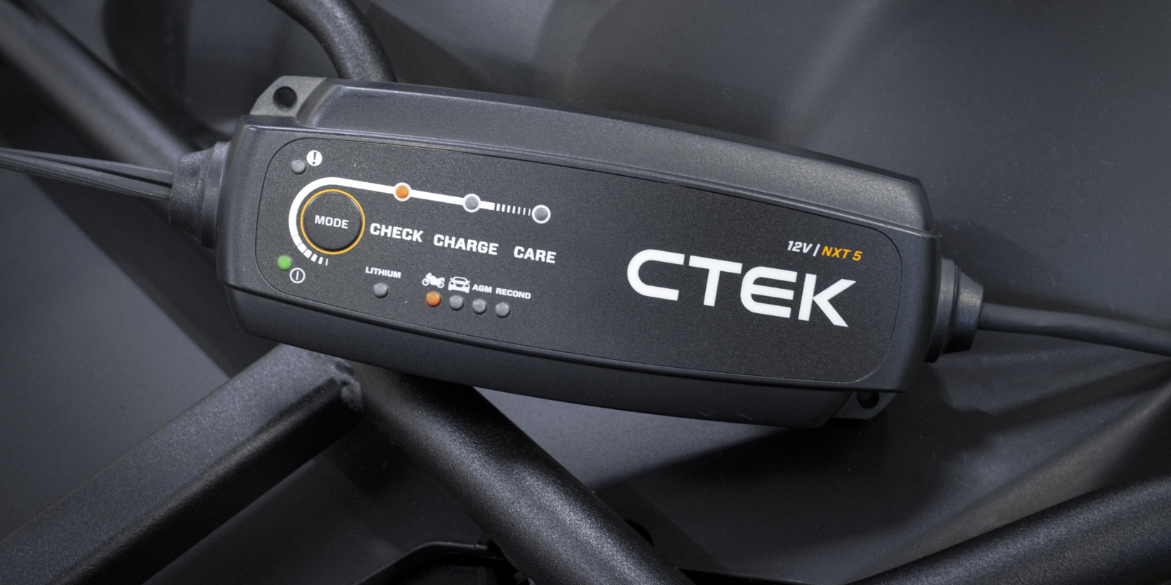 Nærbilde av en CTEK 12V batterilader med indikatorlys og knapper for Modus, Sjekk, Lading og Vedlikehold på en mørk overflate.