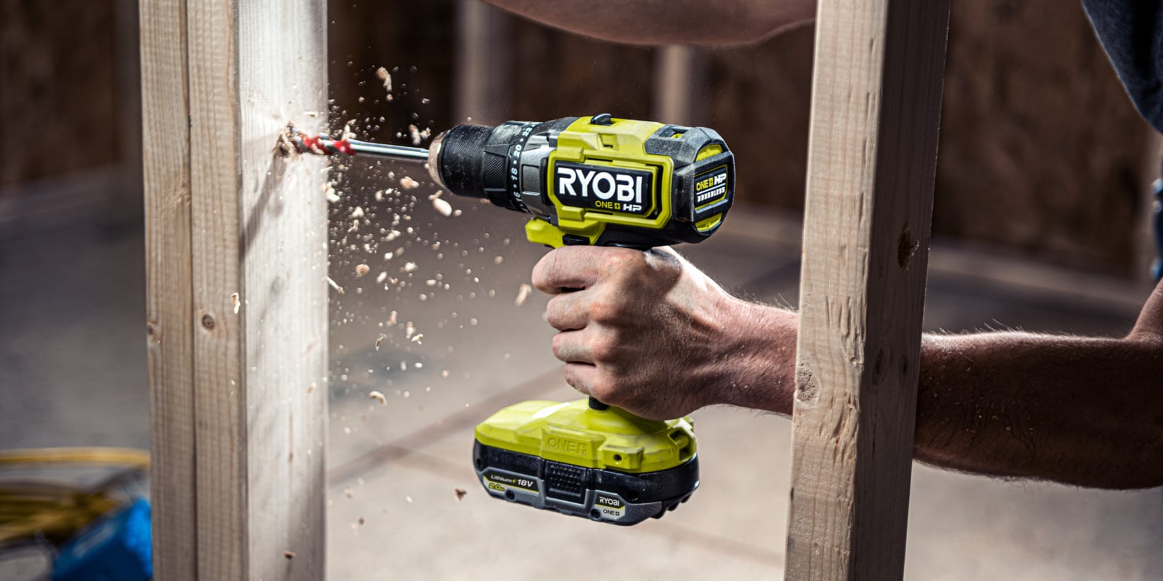 Någon använder en Ryobi sladdlös borrmaskin och borrar ett hål i en träplanka medan träspån sprids från den borrade platsen.