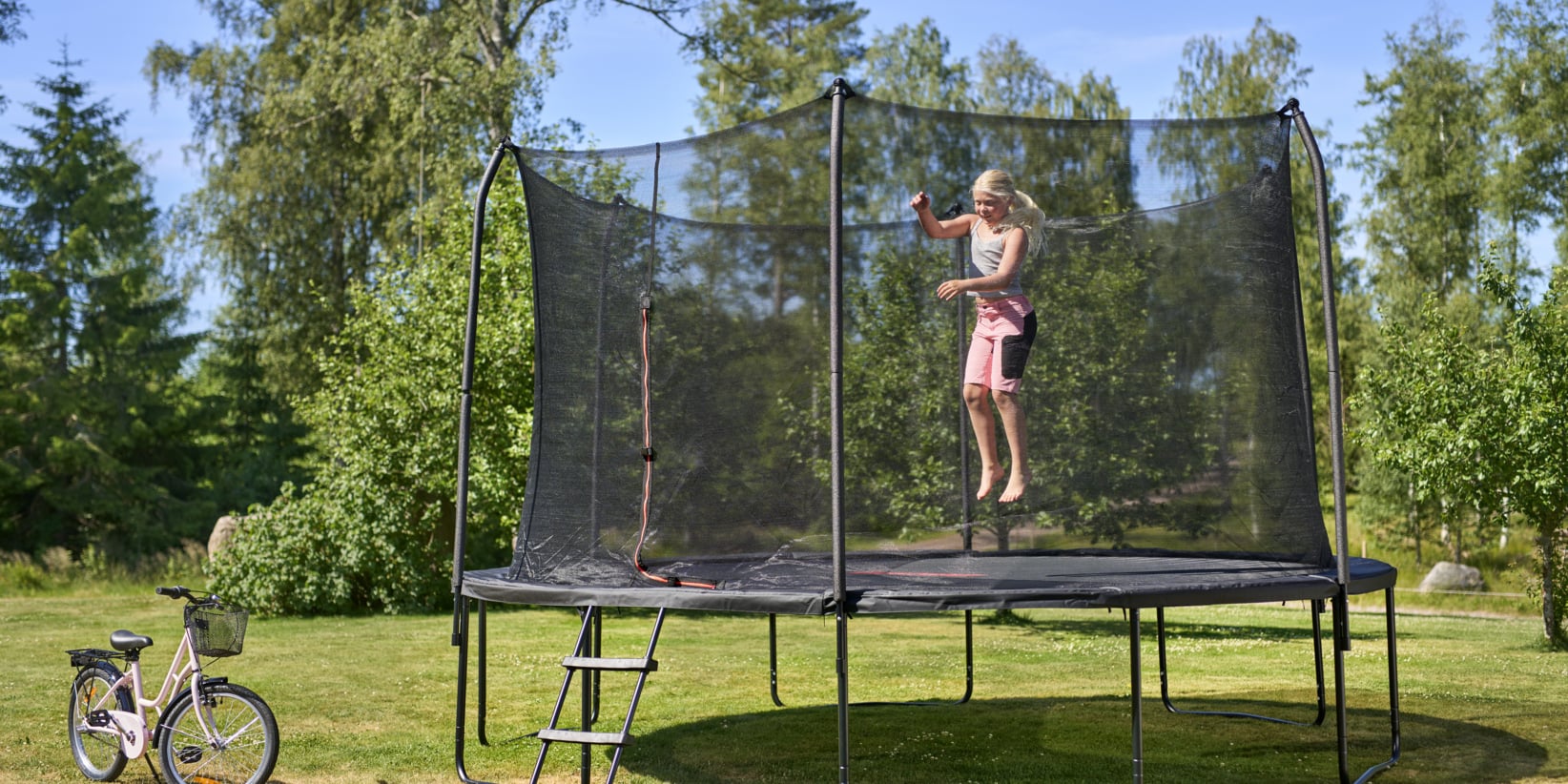 Lapsi raidallisessa paidassa ja vaaleanpunaisissa shortseissa hyppää trampoliinilla nurmikolla. Lähellä on pyörä ja taustalla reunustavat puut.