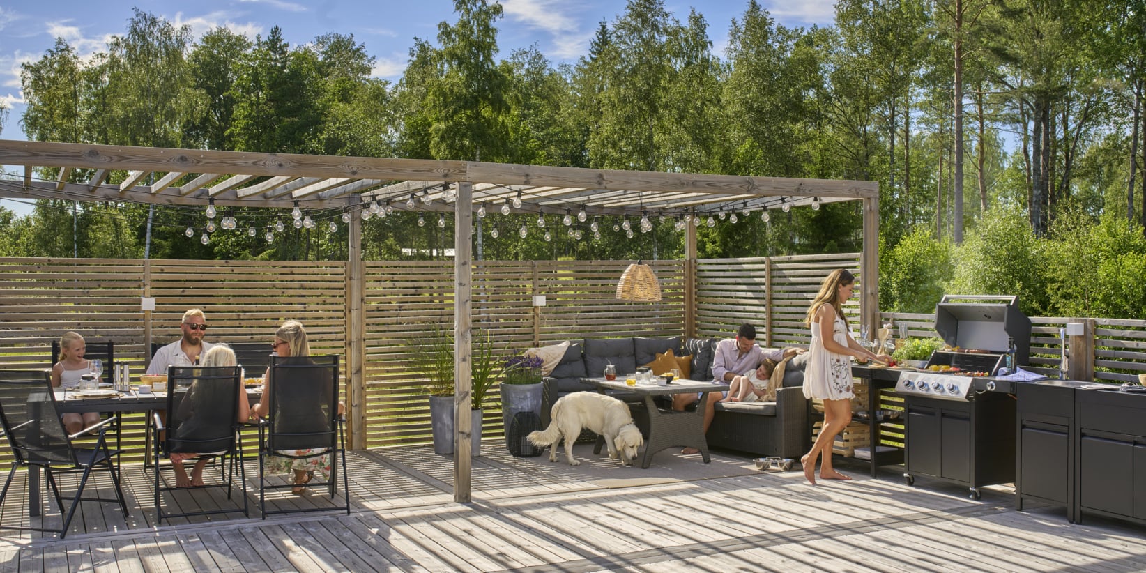 Folk slapper av i en solrik bakgård under en pergola—litt spising, en grilling, en på en sofa med en hvit hund i nærheten. Trær og gjerder omringer.