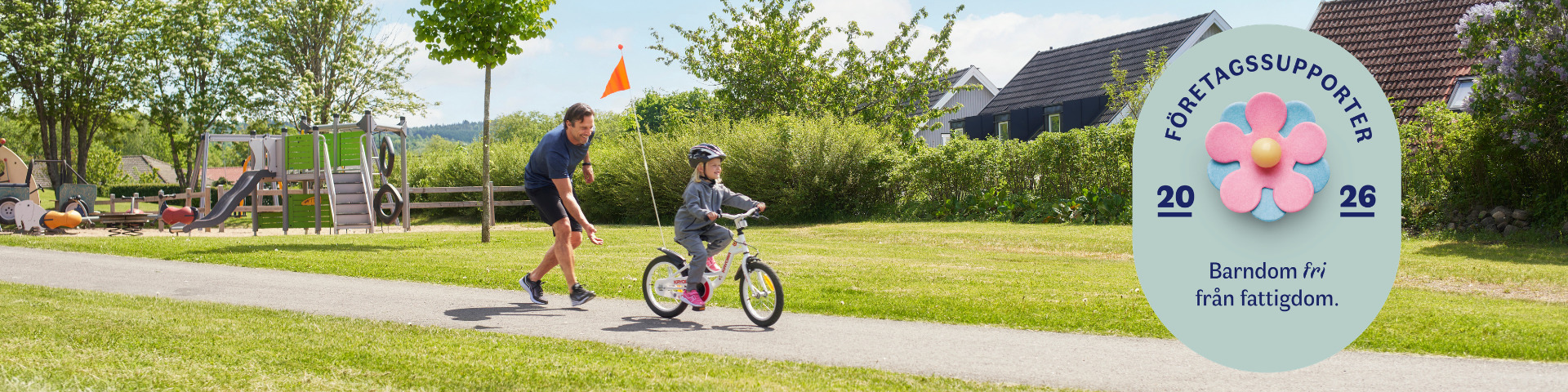 En man hjälper ett barn att cykla på en parkväg. Lekplats, hus i bakgrunden. På badgen står det Företagssupporter 2026 Barndom fri från fattigdom.
