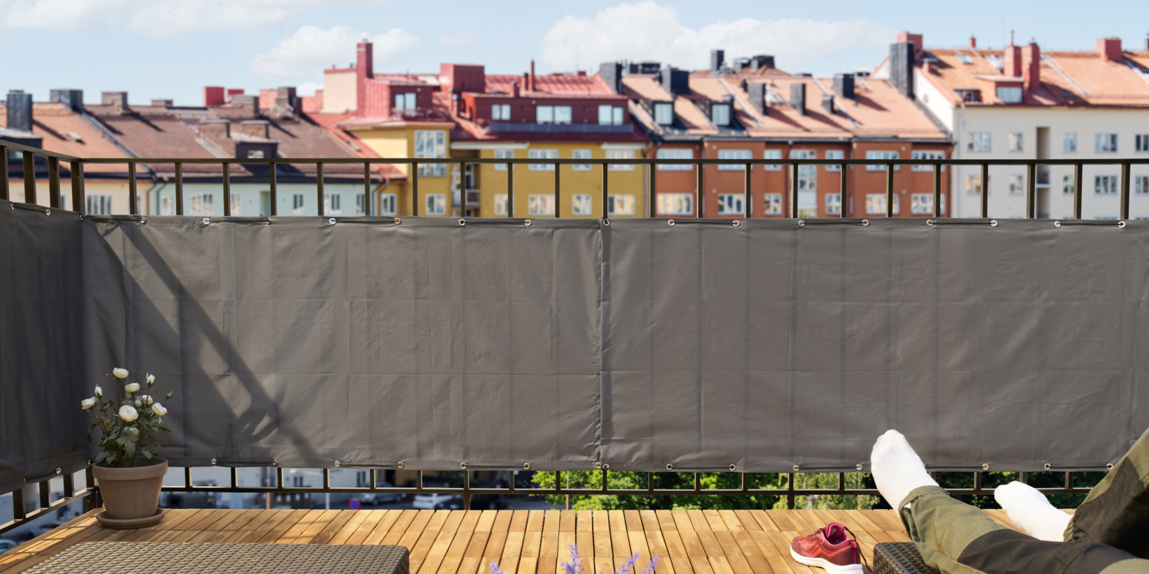 Eine Person, die sich mit hochgelegten Füßen auf einem Holzbalkon entspannt, rote Turnschuhe und eine Topfpflanze in der Nähe, bunte Wohnungen dahinter.
