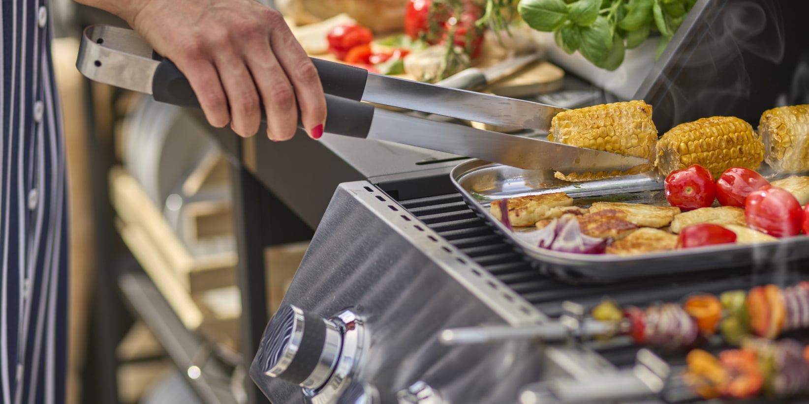 Man grillar majs, tomater och andra grönsaker med tång. Spett med blandade grönsaker och grillade rätter dyker upp framför.