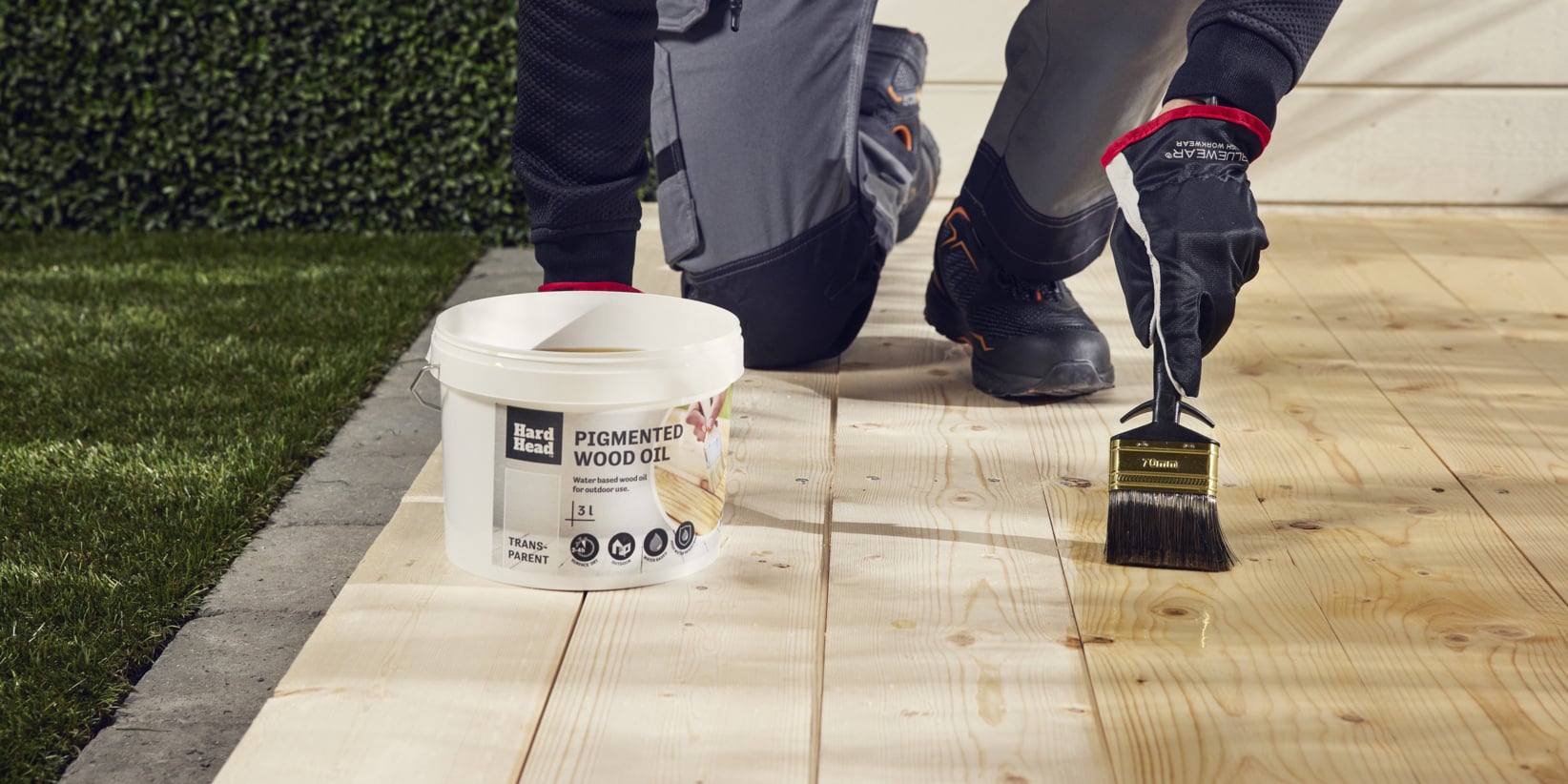 Handskbeklädda händer använder en pensel för att applicera träolja på en altan. En burk märkt Pigmented Wood Oil står bredvid dem; Gräs och asfalt bakom.